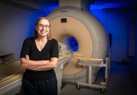 Professor Susan Francis Female, wearing glasses with her arms folded, standing in front of an MRI machine
