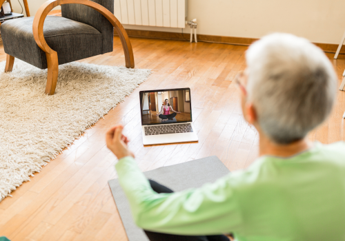 Image - Home Exercise Wellbeing Person sitting on floor crossed legged, with back to the camera. Laptop in front on the floor showing exercises.