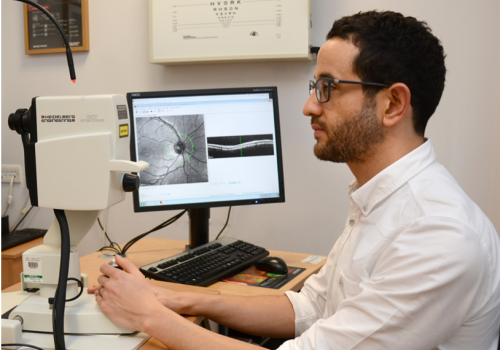 Dr Tariq Farrah Man sat at a desk, with a computer monitor showing which a scan of an eye image.
